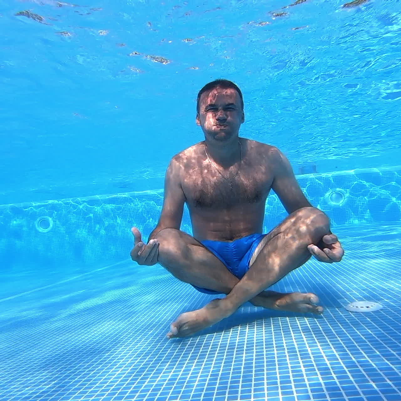 Man sits on the bottom of swimming pool. Healthy man is sitting in yoga pose underwater on blue background. Underwater sport.