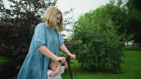 a mother rides a scooter with her son on a peaceful walkway, using her leg to move the scooter forward, the child is smiling with happiness