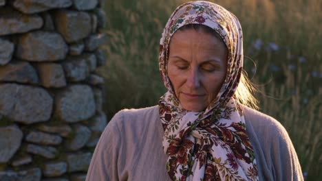 mature woman with closed eyes and headscarf standing serenely in a field at sunset, engaging in prayer or meditation against a backdrop of a stone wall