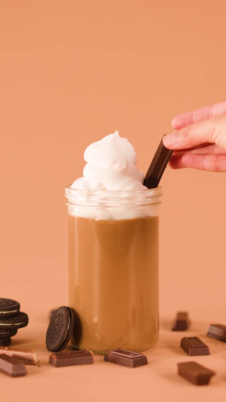 Una mano posiziona una barretta di cioccolato in un milkshake con panna montata, circondato da biscotti e pezzi di cioccolato
