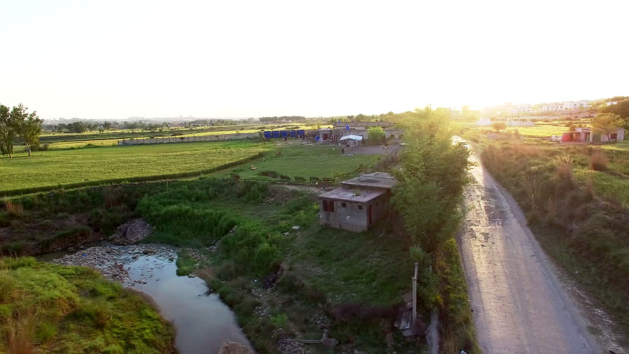 Un rapido sorvolo aereo su una fattoria rigogliosa in controluce e una strada dove circolano auto e biciclette. Il raccolto ondeggia al vento. Un rubinetto per l'acqua di scarico in fattoria
