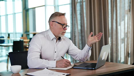 serious businessman looks carefully at his laptop. man stops taking notes into paper notebook and leans back in his chair.