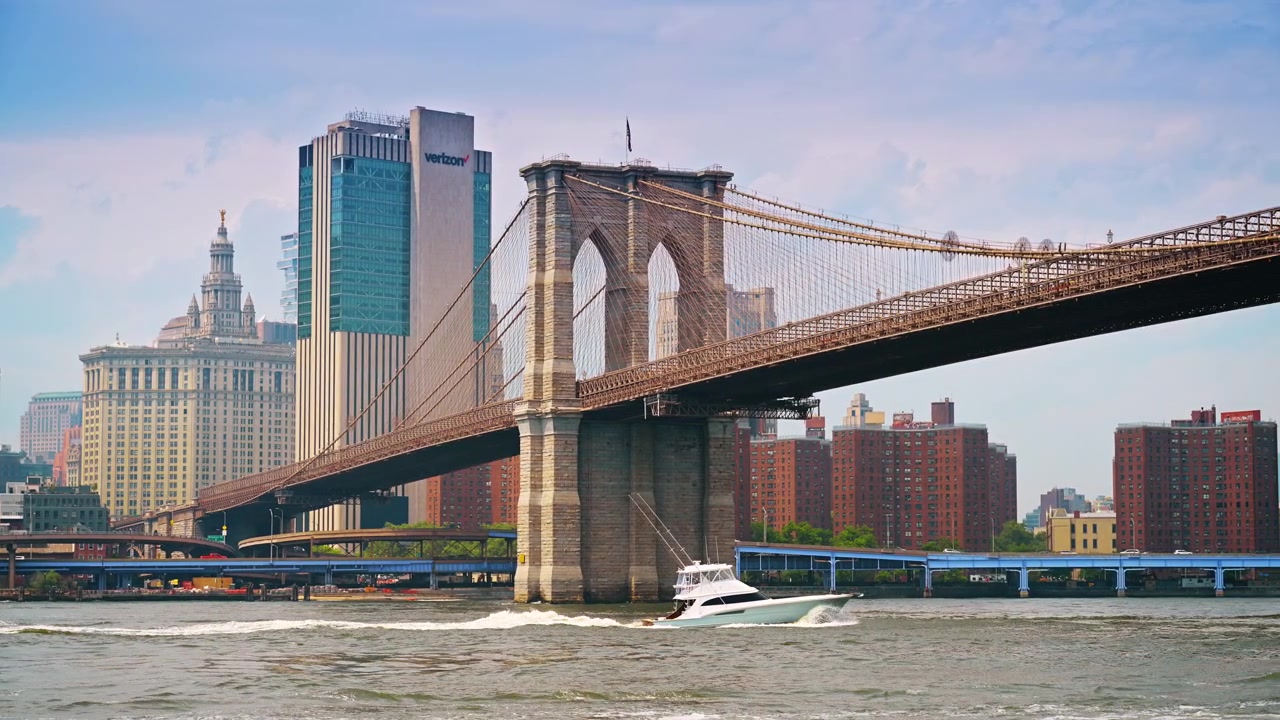 New York, USA, 19 maggio 2026: Vista dal basso della torre del Ponte di Brooklyn e di una barca, New York. Motoscafo che attraversa la superficie dell'acqua accanto all'arco in muratura di granito e agli edifici per uffici
