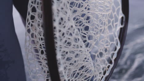 close up of fly fishing net hanging over water beside fisherman legs