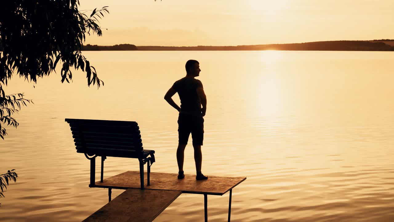 A man was standing on the bridge and than sat down on a bench to looking at the beauty of nature on the background of the river and the sunset on a summer evening.