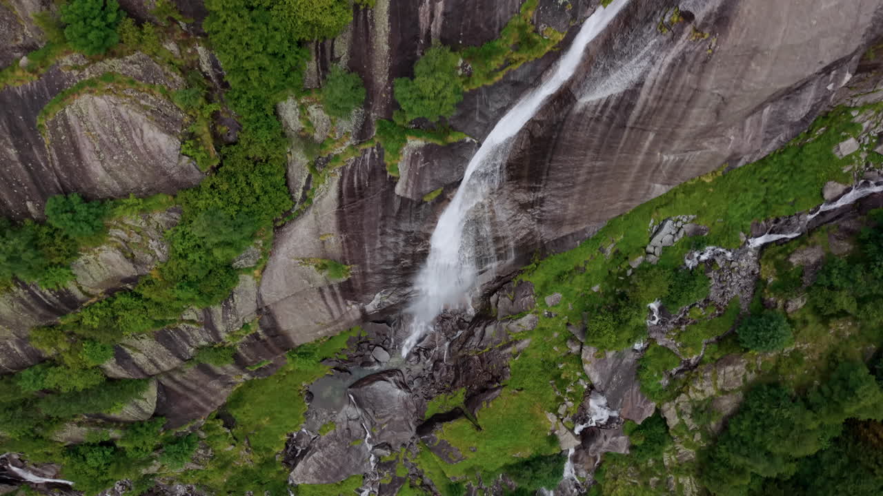 Aerial drone shot in full top-down view flying over a long waterfall on a rocky mountain slope in the Swiss Alps, continuing forward past the falling water