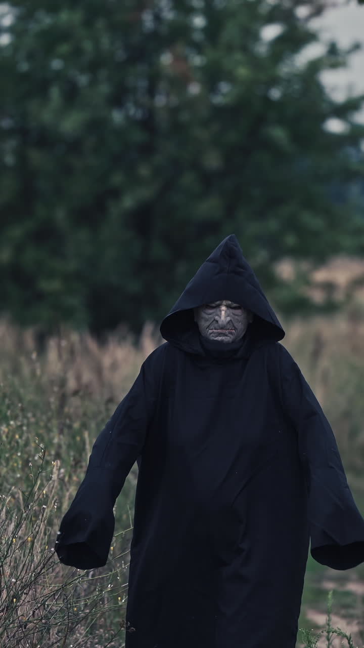 Portrait of scary death in black. Spooky figure in dark mantle with hood coming to camera on nature background. Horror concept. Vertical video