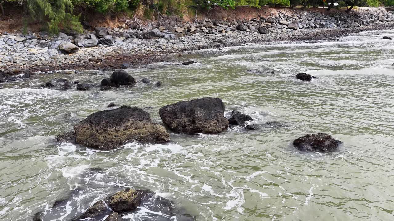 Gentle ocean waves move around large rocks on a rugged, natural shoreline under daylight
