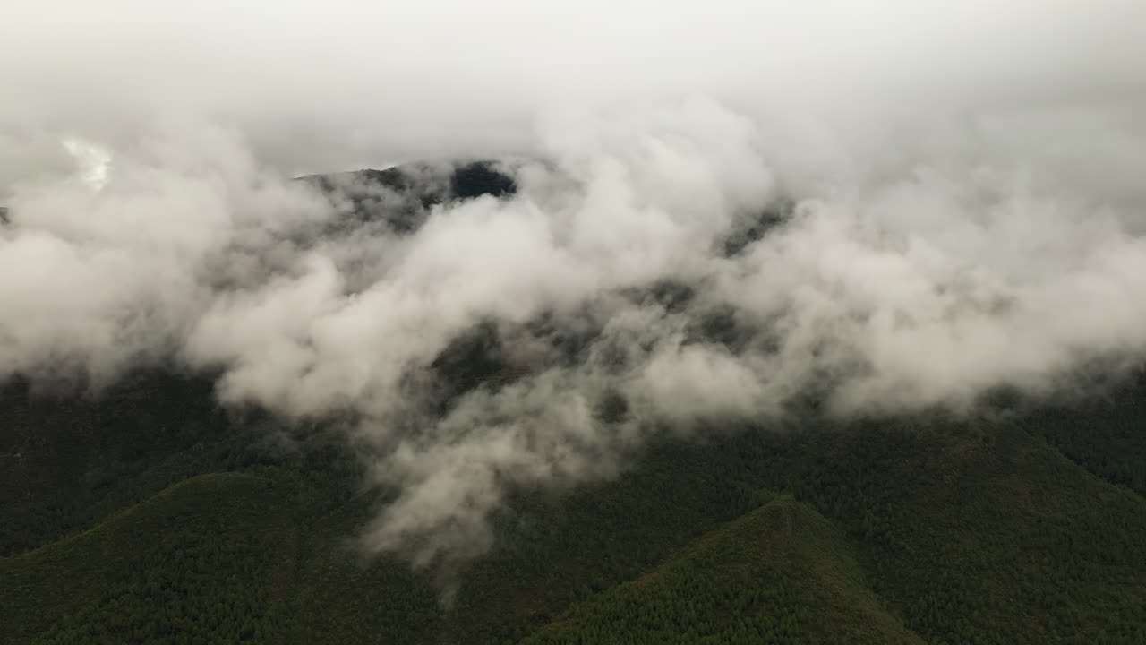 Drone shot of green mountains surrounded by dramatic grey clouds. The camera glides beside the peaks, revealing a moody and atmospheric landscape.