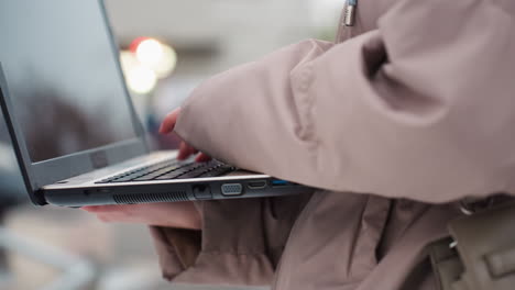 Close-up view of light skin lady typing on laptop while standing outdoors in winter jacket, with blurred urban background and soft light, showing concentration and digital engagement in public space