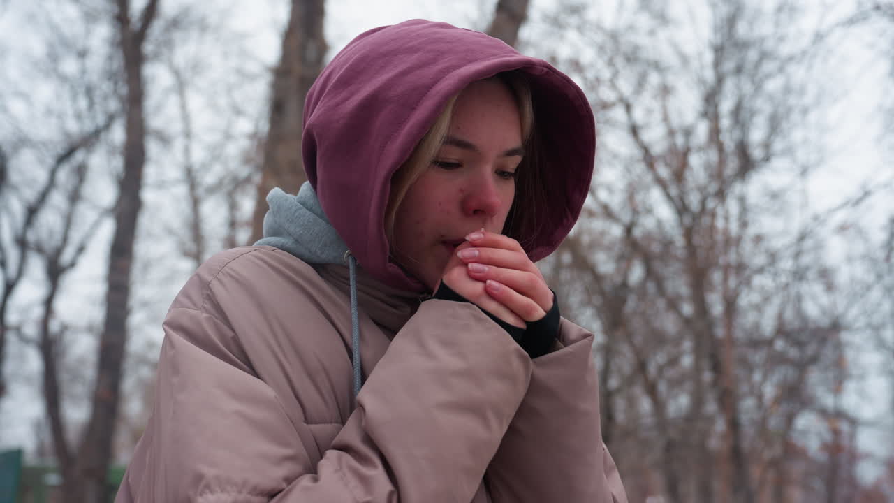 Close-up of a woman feeling cold as she adjusts her hoodie while walking outdoors in winter, she is bundled in a thick jacket, bracing against the chilly air, surrounded by bare trees
