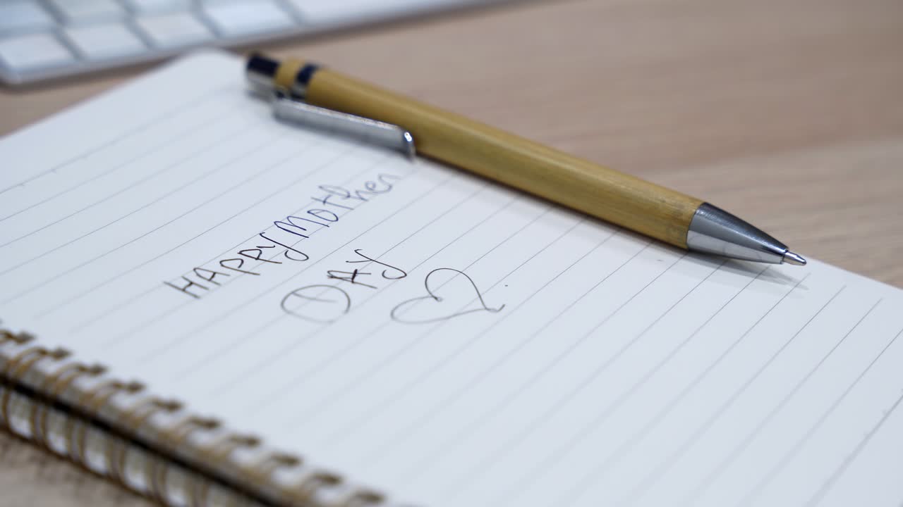Close-up of a spiral notebook with Happy Mothers Day handwritten text and pen on a wooden desk, with blurred keyboard in soft indoor light and shallow depth of field