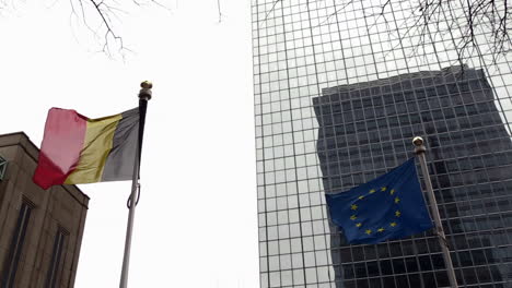 a belgian and european flag waving in the wind on a cloudy day in front of building in brussels