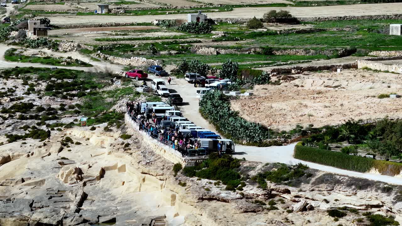 Parlak güneşli bir bahar gününde, Malta'nın Gozo adasındaki engebeli kireçtaşı kayalıkları boyunca park etmiş uzun bir sıra beyaz safari cipini, canlı yeşil tarlalar ve mavi denizi gösteren geniş açılı yüksek bir çekim.