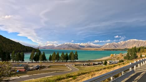 sweeping roadside view of lake tekapo, turquoise water, and snow-capped mountains under dramatic skies, captured in smooth daylight vehicle motion