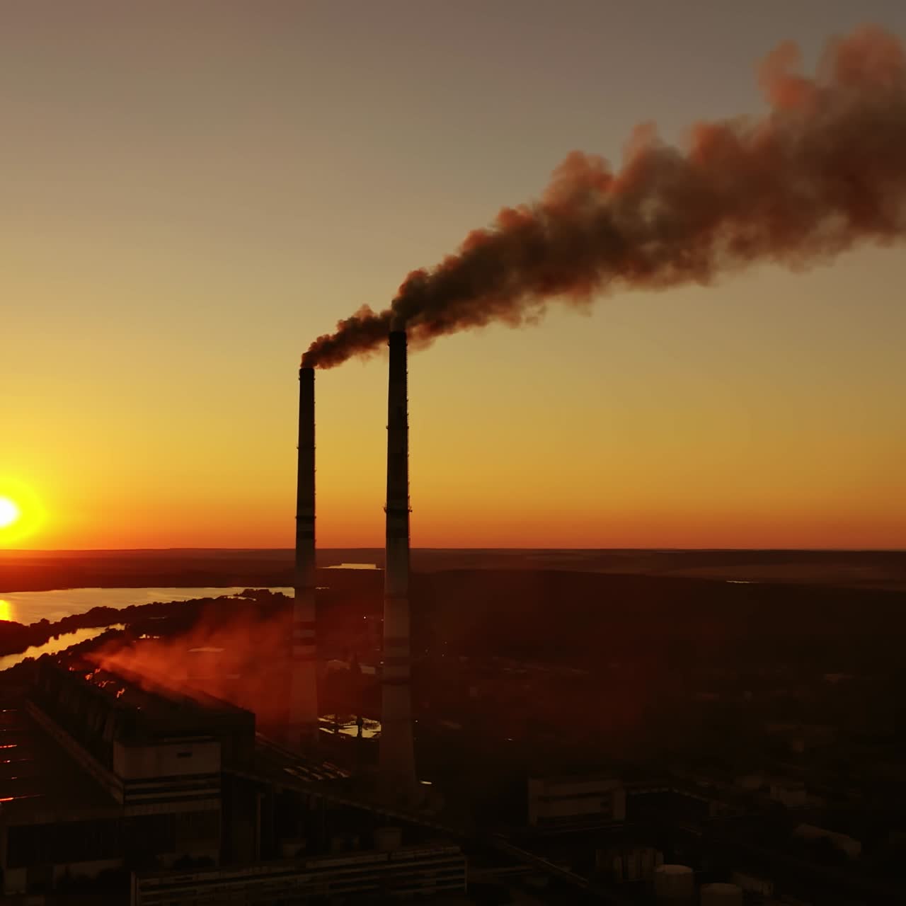 Air pollution. Dark smoke goes from industrial pipes at sunset. Silhouette of harmful factory near the river against the setting sun.