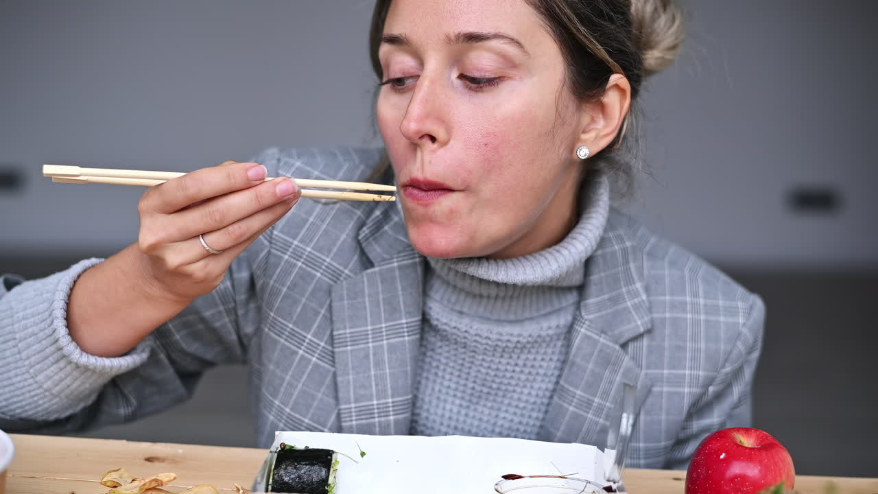 A woman sits at a wooden table, savoring sushi rolls using chopsticks. An apple rests beside her as she focuses on her meal. The setting is casual and cozy