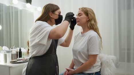 beautician in black gloves applies liquid substance to client eyebrow while seated beside white table filled with makeup tools in well-lit salon with white curtains and vanity light