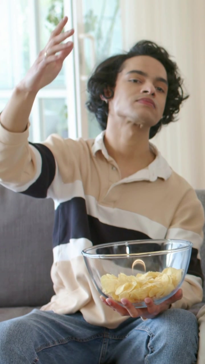 Video verticale: Uomo sul divano che mangia una patatina, facendo un gesto e interagendo fuori campo, mentre tiene una ciotola di vetro piena di patatine.