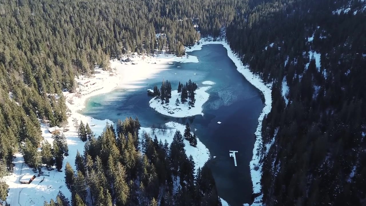 Flygfoto över den delvis frusna schweiziska alpsjön Caumasee, omgiven av en frodig vintergrön skog täckt av snö, belägen i Flims, kantonen Graubünden, Schweiz, under en solig vinterdag.