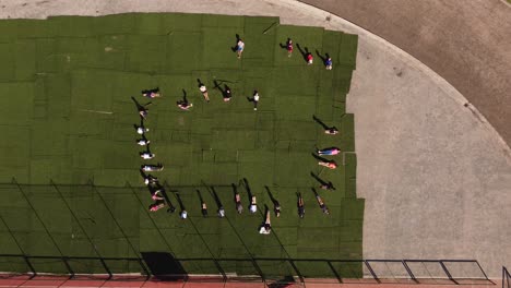 Una-Imagen-Aérea-Descendente-Dinámica-De-Un-Grupo-De-Estudiantes-Tirados-En-El-Césped-Del-Estadio-Atlético-Mientras-Hacen-Ejercicios-De-Elevación-De-Piernas