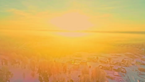 Sobrevuelo-Aéreo-Paisaje-Nevado-De-Invierno-Con-Un-Pequeño-Pueblo-Durante-La-Brillante-Puesta-De-Sol-Dorada-En-El-Horizonte-Iluminando-La-Tierra