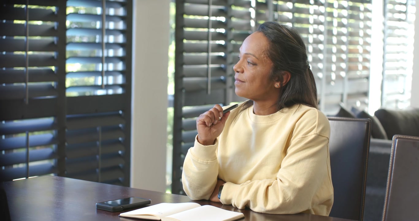 Mature African-American woman writing with pen in journal at table by shutters thinking, copy space