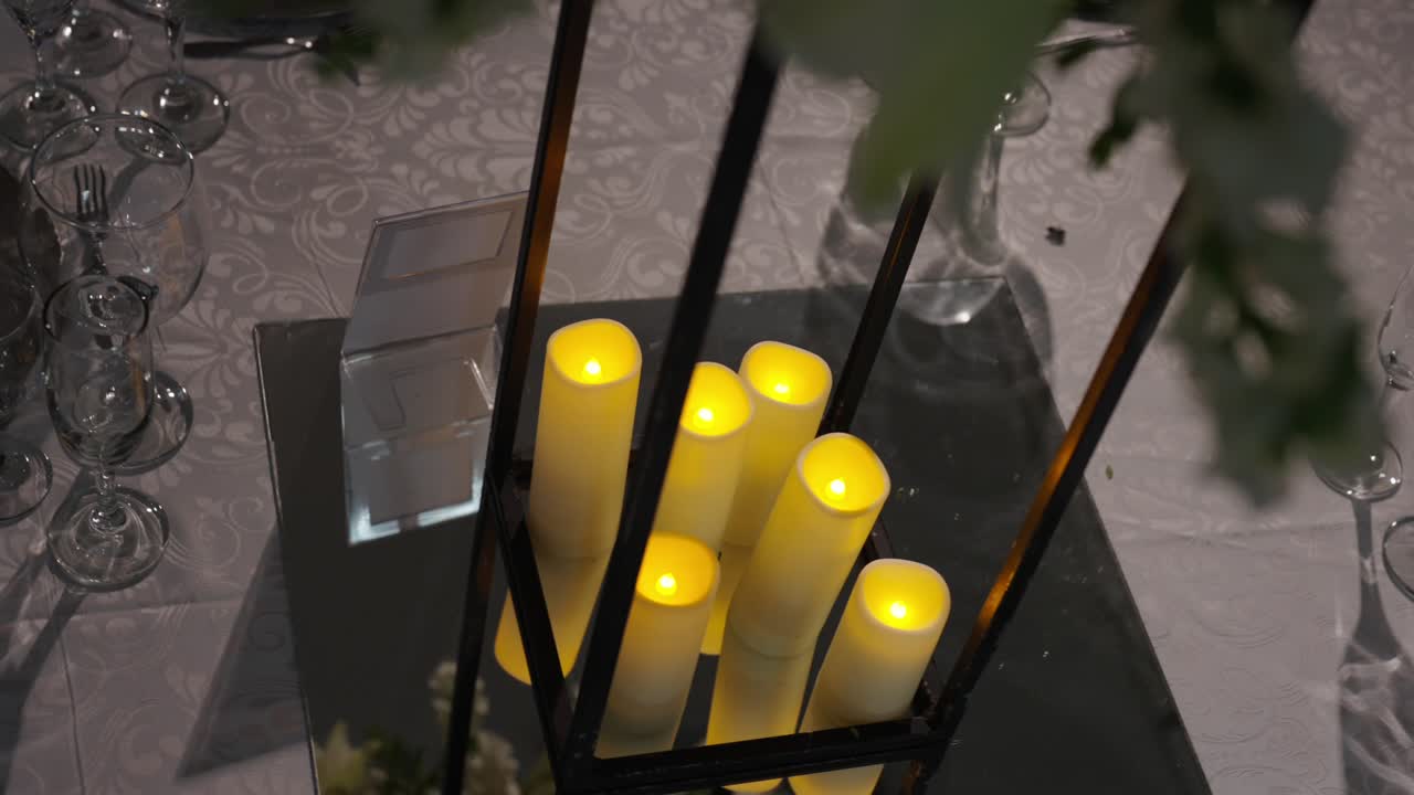 Bröllopsbordsdekoration med LED-ljus inuti glaslykta på reflekterande yta omgiven av glasvaror och tygdukning i inomhuslokal, varm ljusatmosfär, långsam avslöjande omloppsbild.
