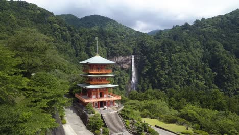 Seigantoji-Temple-from-the-sky,-aerial-flyover-approaching-Nachi-Falls-surrounded-by-a-deep-green-verdant-forest