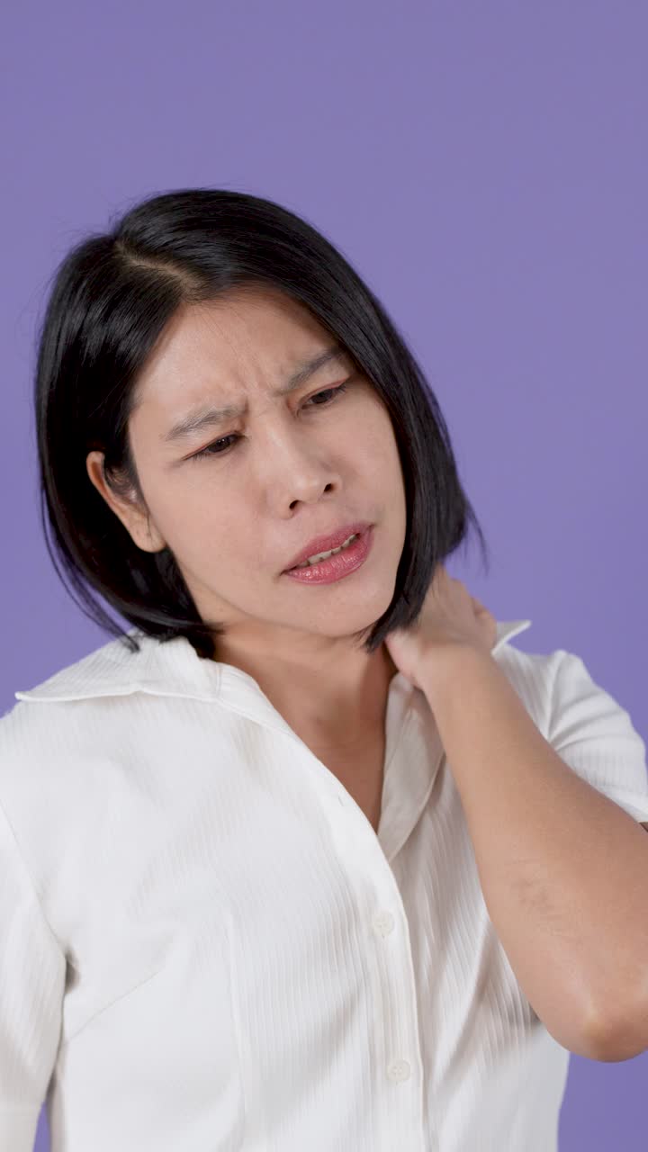Donna asiatica si massaggia il collo e la parte superiore della schiena doloranti, esprimendo il disagio della sindrome da ufficio su sfondo viola.