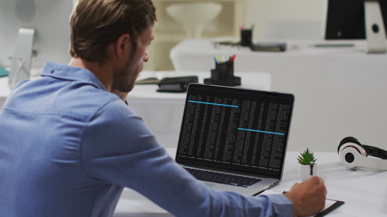 hombre caucásico sentado en un escritorio viendo el procesamiento de datos de codificación en una computadora portátil y tomando notas