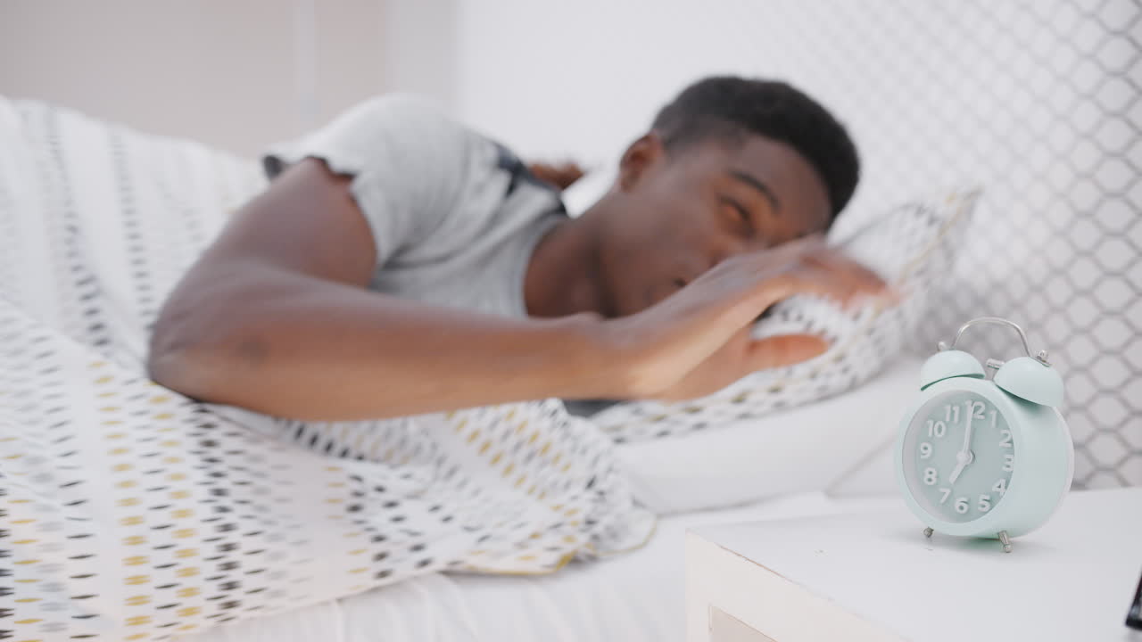 Uomo addormentato a letto si allunga per spegnere la sveglia sul comodino.
