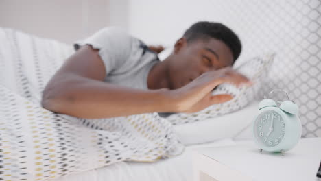 un hombre dormido en la cama extiende la mano para apagar el despertador en la mesa de noche.