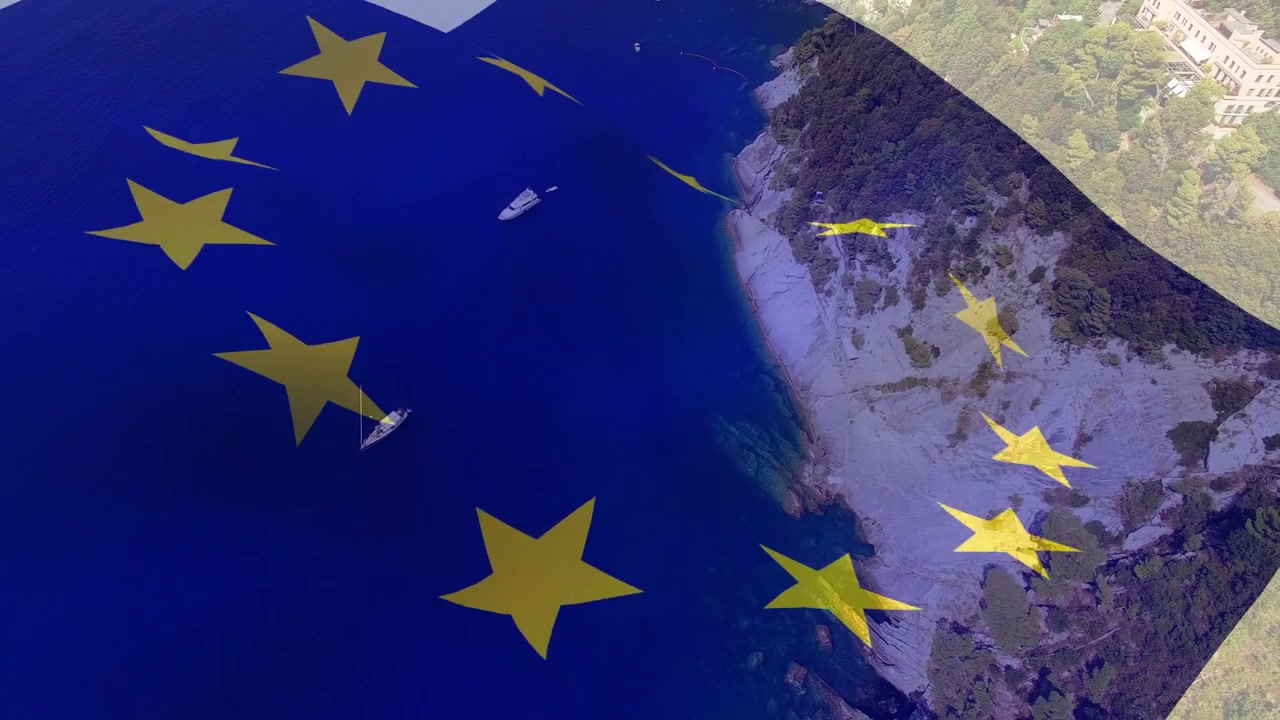 Sovrapposizione della bandiera dell'UE che appare, sventolando su una baia, con le stelle che si spostano rivelando la costa, a significare unità.