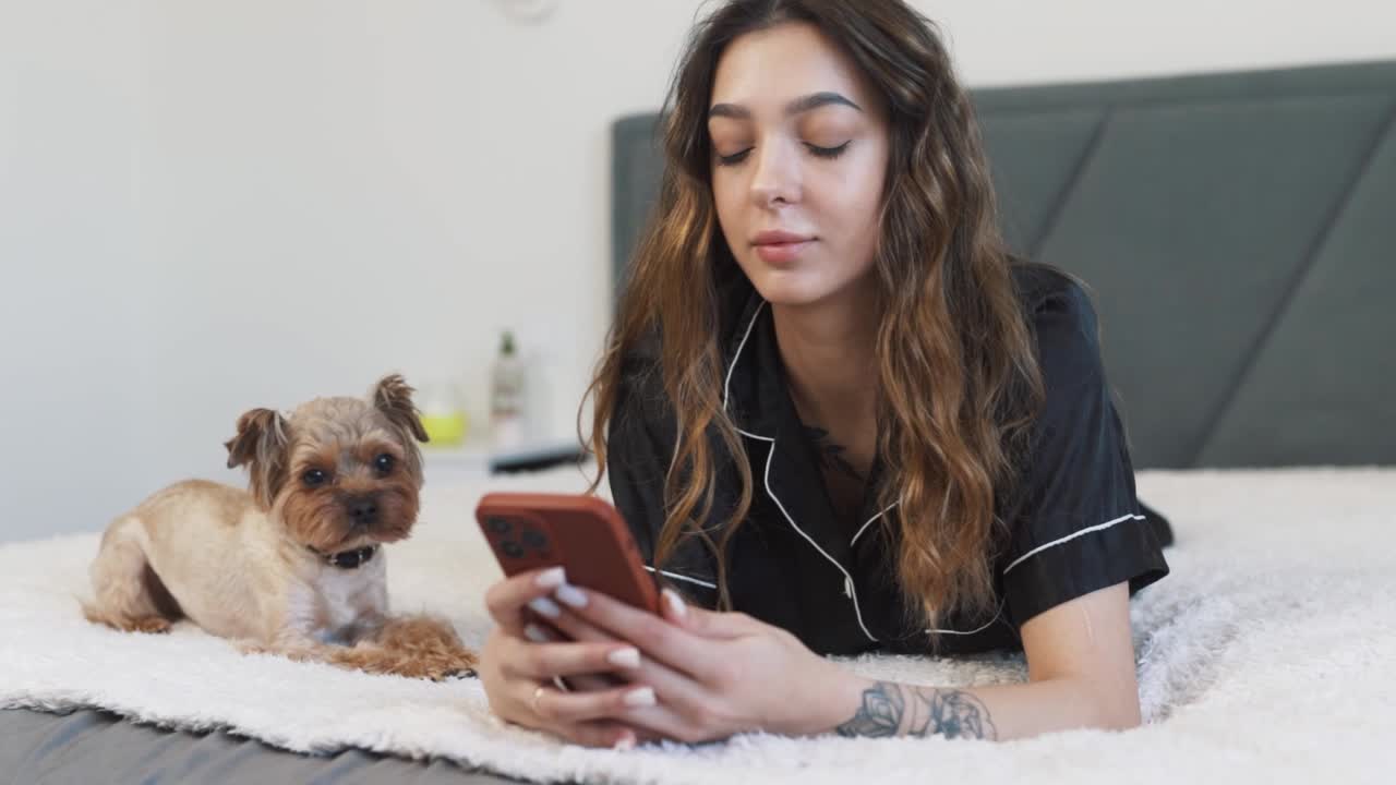 hermosa mujer joven, vestida con una túnica, yace en la cama con un yorkshire terrier, hablando en su teléfono inteligente y sonriendo