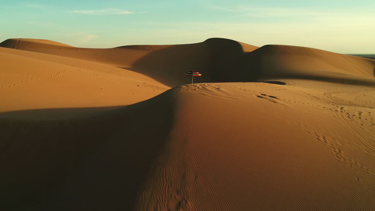 Aerial fly by of colorful beach umbrella isolated in desert sand dunes at sunset