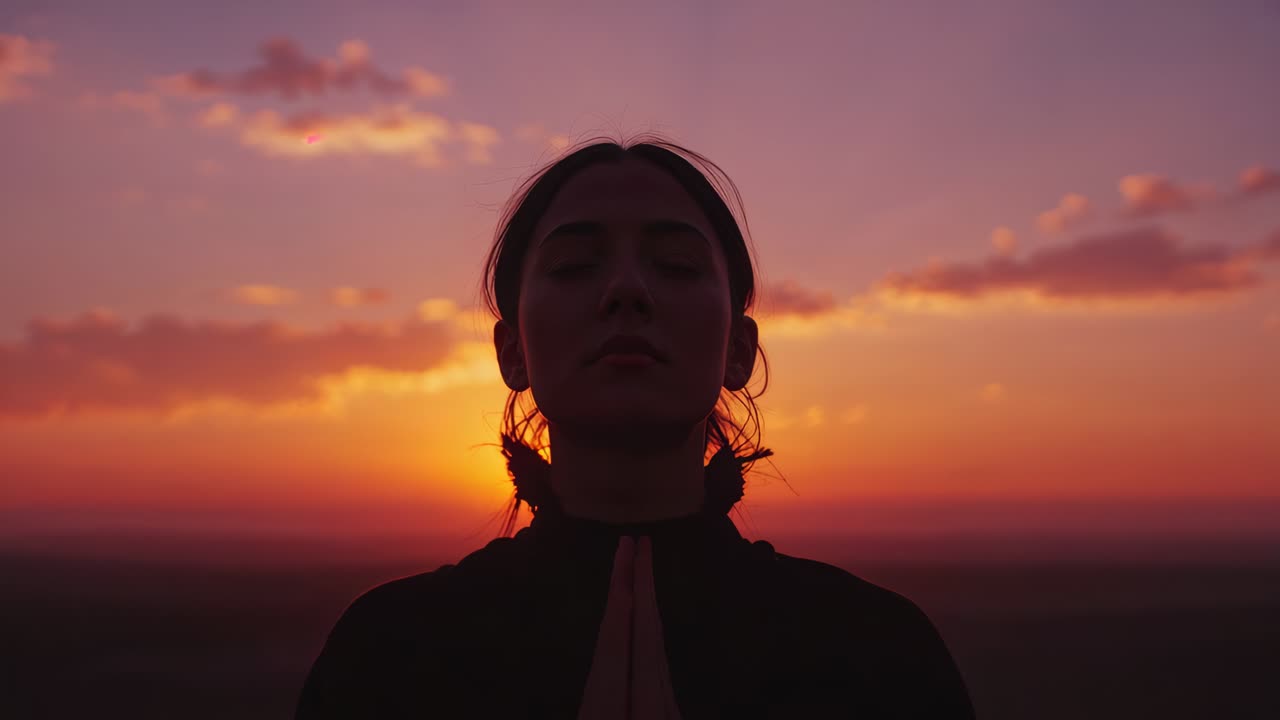 Sun glowing behind woman in dark collared top praying with hands pressed on hilltop revealing smile