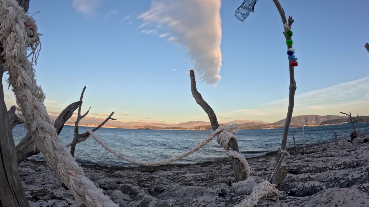 Scena nautica rustica con corde bianche annodate attorno a una struttura di legno spiaggiato che incornicia una vista panoramica sul mare calmo, montagne e una particolare formazione di nuvole al tramonto su una costa rocciosa