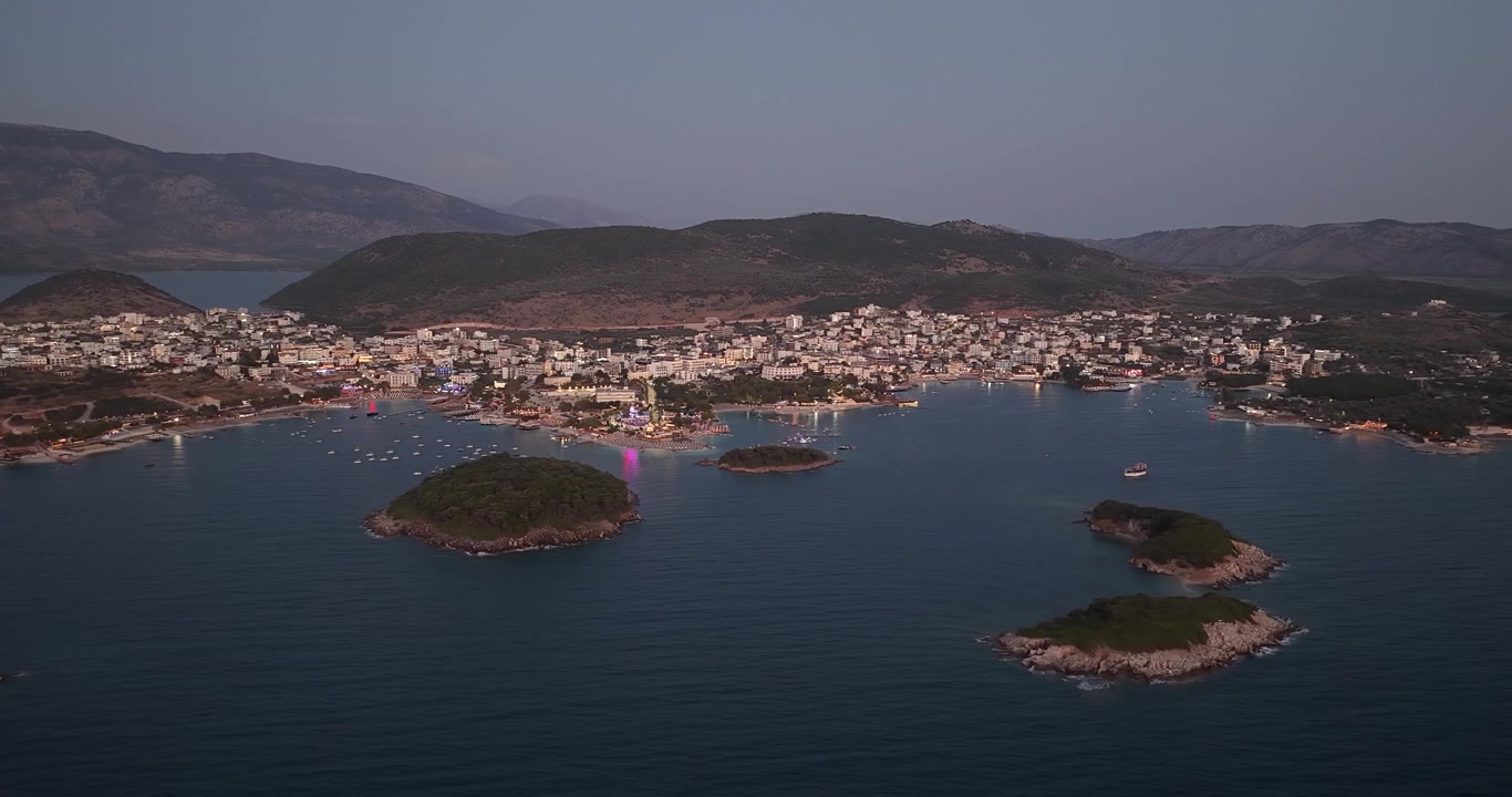 Ksamil, Arnavutluk'un güzel gece ve alacakaranlık drone görüntüsü. Sahil tatil kasabası, koyu mavi denizin ve küçük adaların kenarında parlayan sokaklar ve lunapark ile aydınlanıyor