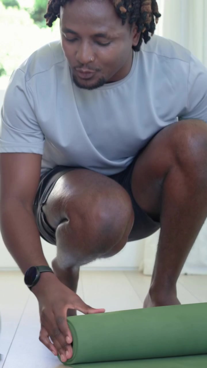 Vertical video: Unrolling man in grey tee aligning mat at home, rising, carrying mat for workout