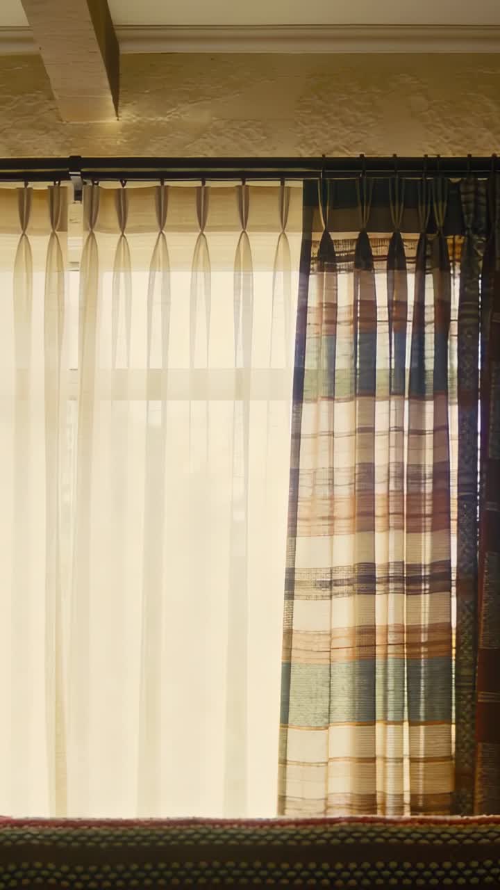 Vídeo vertical: Luz solar em movimento banhando janela com montantes em casa, movendo cortina transparente estampada.