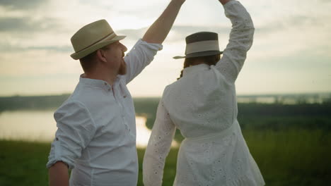 close up as a man spins his wife around on a grassy hill beside a peaceful lake at sunset. the man, dressed in a white shirt, hat, and jeans, as the woman in a white dress and black hat embraces him