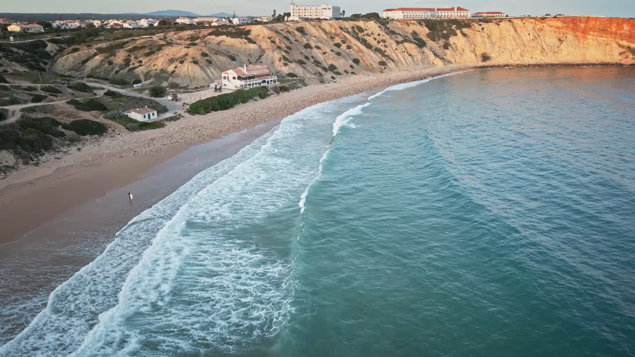 Moving forward from turquoise water to sandy beach and golden cliffs, panning up to unveil the scenic coastline and coastal buildings near Sagres, Portugal