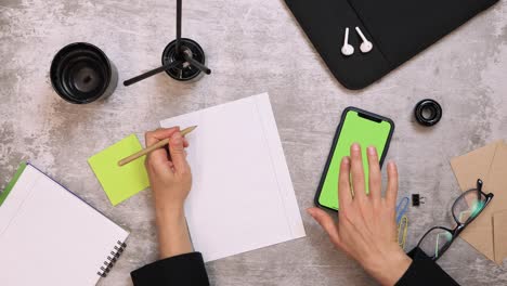 woman writing notes with pen on paper, top view of office work desk. female sits at table with smartphone with green screen chrome key. woman sitting at desk in office, writing necessary information.