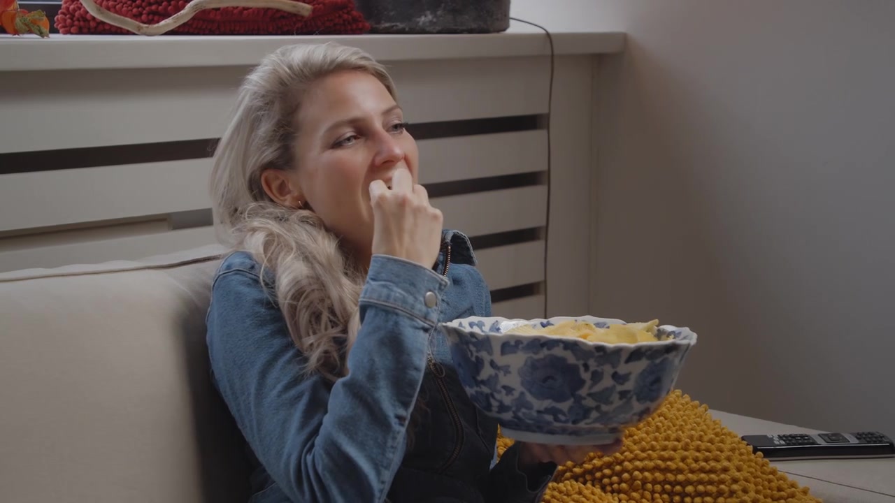 Joven mujer caucásica sentada en un cómodo sofá, navegando por los canales y viendo algo aburrido o frustrante mientras come de un tazón de patatas fritas