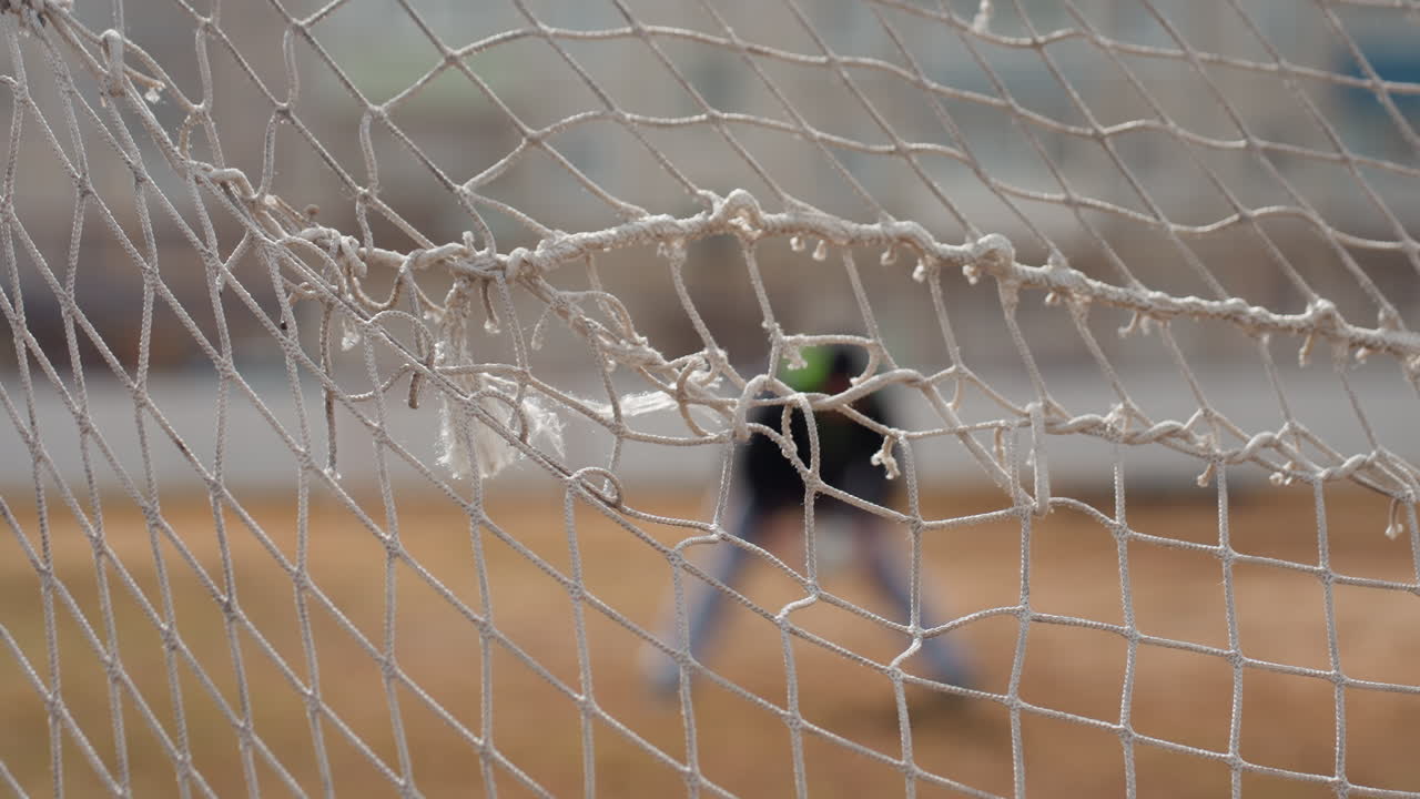 close-up view net with ball entangled and blurred goalkeeper in background, caucasian player silhouette and stadium fence visible, nylon mesh texture and tension of score moment on urban pitch