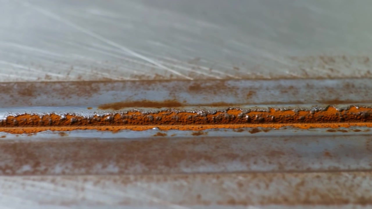 Close view of rust spreading along horizontal weld seam on corrugated metal panel, showing granular oxidation, flaking edges, pitting, muted colors, worn industrial texture, and weathered surface