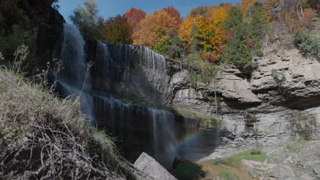 a scenic waterfall with a rainbow below, surrounded by vibrant autumn foliage and rocky cliffs