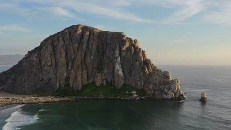 low-altitude-aerial-drone-circling-Morro-Bay-Rock-Beach-during-sunset-in-California-USA-as-waves-come-crashing-onto-the-beach