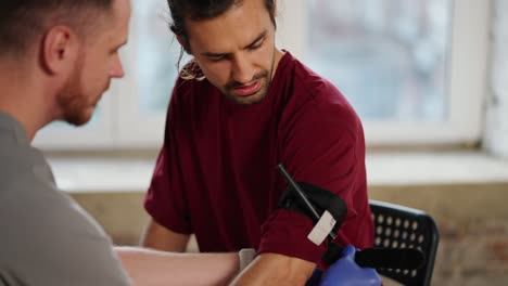 Close-up-shooting:-A-bearded-brunette-man-in-a-gray-T-shirt-tightens-a-tourniquet-with-another-bearded-brunette-man-with-long-hair-in-a-red-T-shirt.-Training-of-practical-classes-of-first-aid